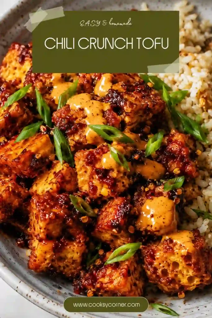 A bowl of crispy Chili Crunch Tofu served over steamed rice and topped with sliced green onions. The baked tofu is coated in a creamy spicy chili crisp sauce with a golden, crispy exterior.