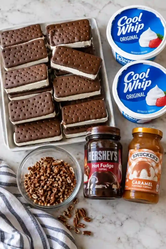 Easy Ice Cream Sandwich Cake topped with whipped topping, glossy chocolate fudge, golden caramel, and crunchy chopped pecans. Visible layers of vanilla ice cream sandwiches create a creamy no-bake frozen dessert.